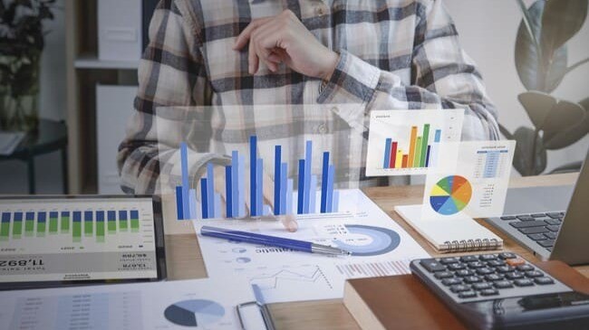 Pessoa analisando gráficos financeiros com tablets, papéis, calculadora e laptop sobre a mesa.